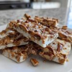 Stack of homemade peanut butter pretzel bark pieces topped with sea salt on a white plate.