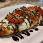 A close-up of a grilled caprese chicken breast topped with melted mozzarella, sliced tomatoes, fresh basil, and balsamic glaze.