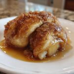 A single, glistening apple dumpling covered in cinnamon sugar glaze, sitting in a pool of sauce on a white plate.
