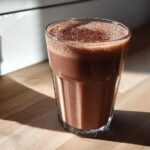 A tall glass filled with rich, frothy chocolate milk, dusted with cocoa powder, sitting on a wooden counter.