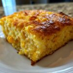 A close-up of a moist slice of cornbread casserole with visible corn kernels, served on a white plate.