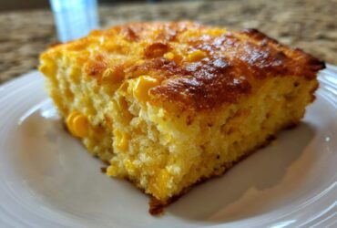 A close-up of a moist slice of cornbread casserole with visible corn kernels, served on a white plate.