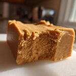 A thick, square piece of creamy peanut butter fudge resting on a white cutting board, showing a bite taken out.