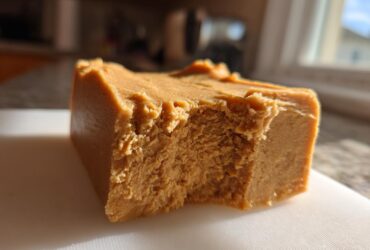 A thick, square piece of creamy peanut butter fudge resting on a white cutting board, showing a bite taken out.