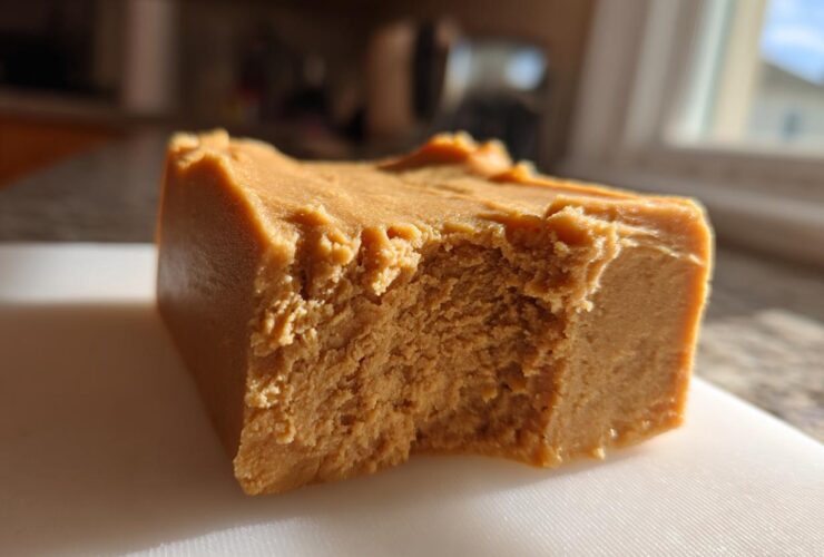 A thick, square piece of creamy peanut butter fudge resting on a white cutting board, showing a bite taken out.