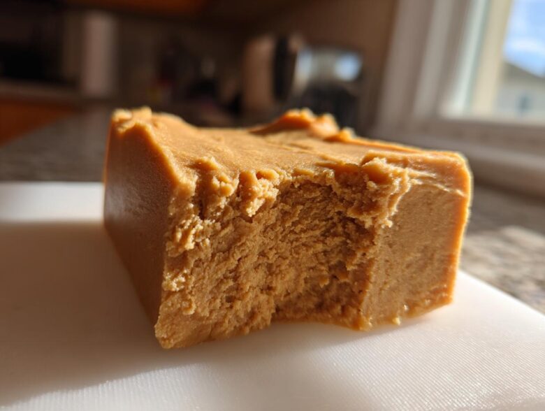 A thick, square piece of creamy peanut butter fudge resting on a white cutting board, showing a bite taken out.