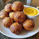 A plate of freshly baked, golden brown pretzel bites sprinkled with coarse salt, served with a side of bright yellow cheese dip.