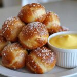 A pile of golden brown pretzel bites sprinkled with coarse salt next to a white ramekin of bright yellow cheese sauce.