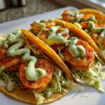 Three delicious shrimp taco shells filled with shredded lettuce, seasoned shrimp, and drizzled with a creamy green sauce.