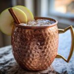 A frosty apple cider moscow mule served in a hammered copper mug, garnished with green apple slices and a cinnamon stick.