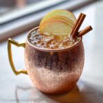 A condensation-covered copper mug filled with an apple cider Moscow mule, garnished with apple slices and cinnamon sticks.