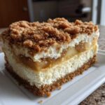 A thick, layered slice of apple pie cheesecake topped with cinnamon crumble, sitting on a white plate.