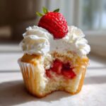 A close-up of a strawberry shortcake cupcake with a bite taken out, revealing strawberry filling, topped with whipped cream and a fresh strawberry.