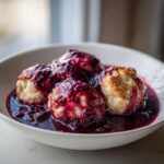 Four fluffy Blackberry Dumplings served warm in a white bowl, generously covered with a dark, rich blackberry sauce.