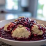 Close-up of fluffy Blackberry Dumplings swimming in a rich, dark purple berry sauce in a white bowl.