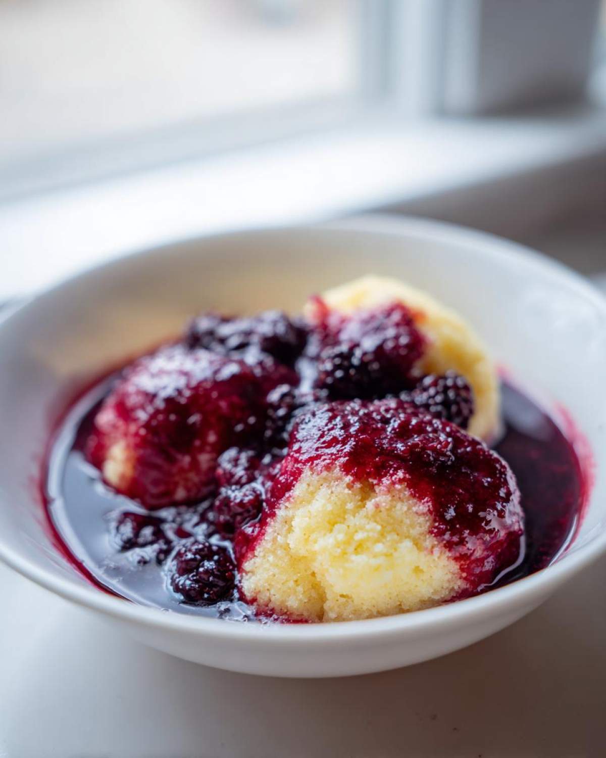 Close-up of fluffy yellow Blackberry Dumplings soaking in rich, dark blackberry sauce in a white bowl.