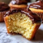 Close-up of a boston cream pie cupcake cut in half showing vanilla cake, pastry cream filling, and chocolate ganache topping.