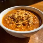 A close-up of a white bowl filled with rich, orange buffalo chicken chili, topped with shredded chicken and beans.