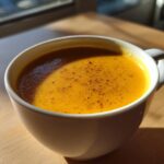 A vibrant orange cup of creamy butternut squash soup, sprinkled with pepper, sitting on a wooden table in sunlight.