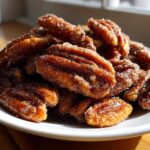 A close-up pile of freshly made candied pecans recipe nuts coated in sparkling sugar, served on a white plate.