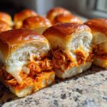 Close-up of two cheesy buffalo chicken sliders with shredded orange chicken filling spilling out of soft, golden buns.