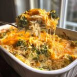 A spoonful of creamy Chicken Divan with shredded chicken, broccoli, and melted cheddar cheese being lifted from the baking dish.
