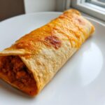 A single baked cheesy pocket tacos, filled with seasoned ground meat, resting on a white plate near a window.