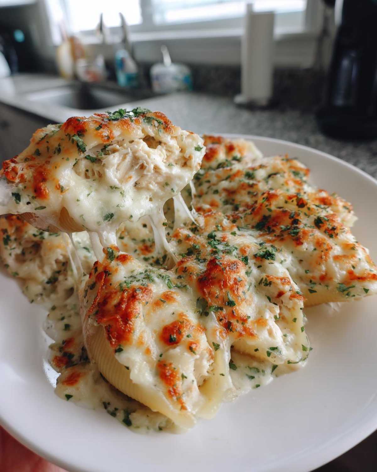 A serving spoon lifts one of the baked chicken alfredo stuffed shells, showing a dramatic cheese pull.