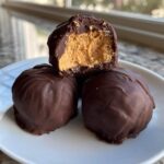 Close-up of three chocolate covered peanut butter balls on a white plate, one is cut in half showing the creamy filling.