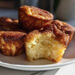 A stack of golden brown french toast muffins coated in cinnamon sugar, with one muffin showing a bite taken out.