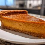 A thick slice of classic pumpkin pie recipe with a rich orange filling and a visible graham cracker crust, served on a white plate.