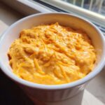 Close-up of a white bowl filled with thick, orange beer cheese dip topped with shredded cheddar.