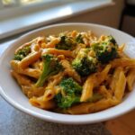 Close-up of a white bowl filled with creamy, orange-sauced broccoli pasta (penne) next to a bright window.