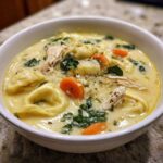 Close-up of a white bowl filled with creamy chicken tortellini soup, featuring tortellini, shredded chicken, carrots, and spinach.