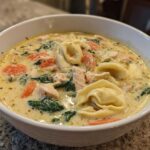 A close-up of a white bowl filled with creamy chicken tortellini soup featuring tortellini pasta, chicken pieces, spinach, and carrots.