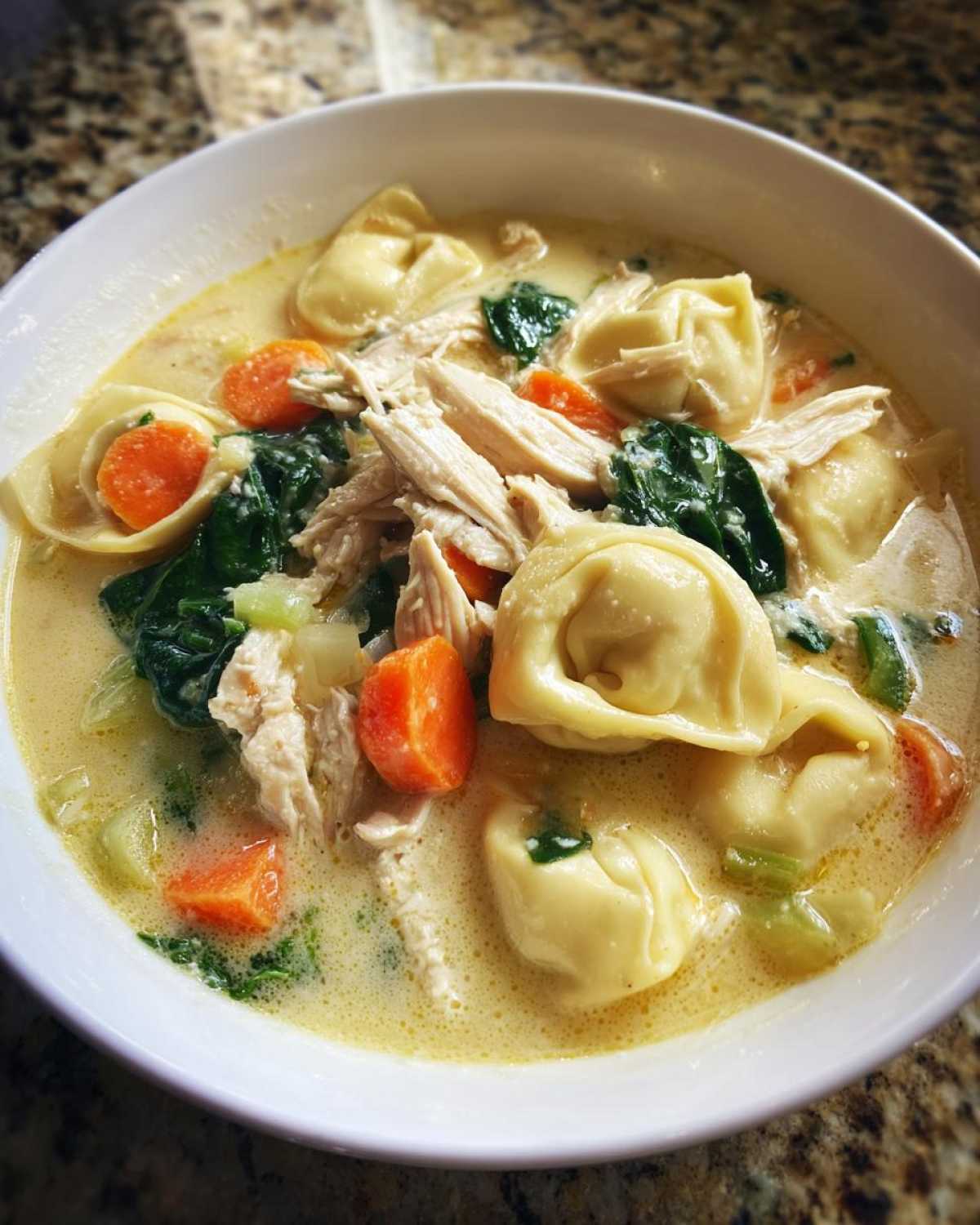 A close-up of a white bowl filled with creamy chicken tortellini soup, featuring tortellini, shredded chicken, carrots, and spinach.