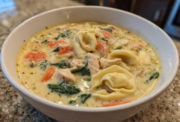 A close-up of a white bowl filled with creamy chicken tortellini soup featuring tortellini pasta, chicken pieces, spinach, and carrots.