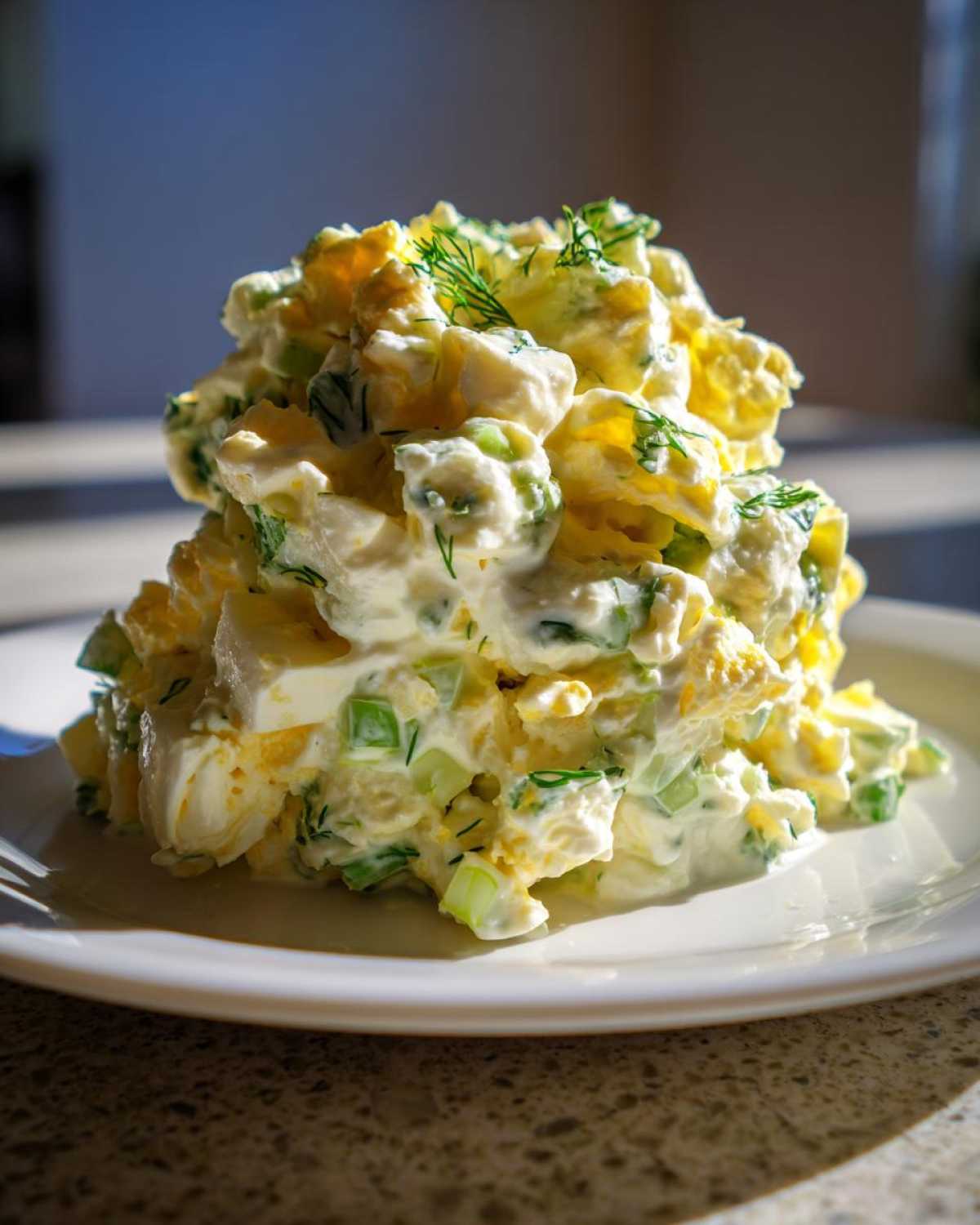 A tall mound of creamy egg salad recipe mixed with celery and dill, served on a white plate.