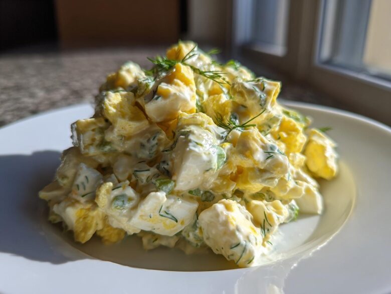 A generous mound of creamy egg salad recipe, featuring chopped eggs, mayonnaise dressing, and fresh dill, served on a white plate.