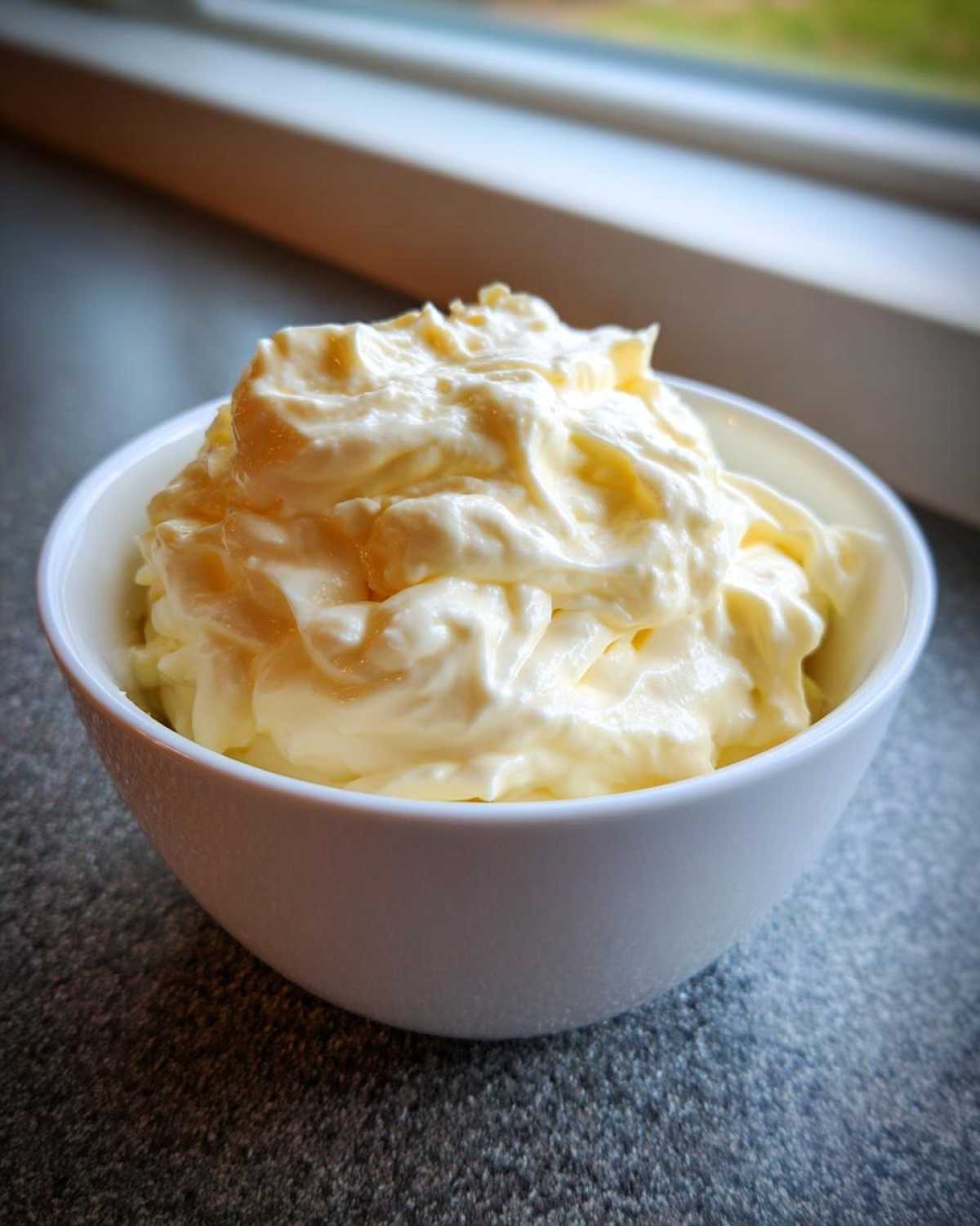 A white bowl filled with thick, pale yellow homemade mayonnaise, set against a dark countertop near a window.