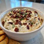 A close-up of a white bowl filled with creamy Neiman Marcus dip, topped generously with bacon bits, sliced almonds, and chives.