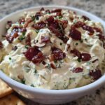 A close-up of a bowl filled with creamy Neiman Marcus dip, topped generously with crispy bacon pieces and chopped chives.
