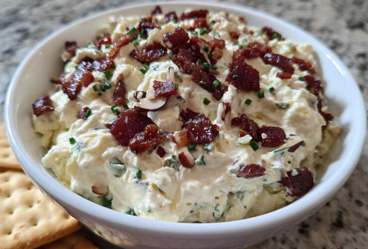 A close-up of a bowl filled with creamy Neiman Marcus dip, topped generously with crispy bacon pieces and chopped chives.