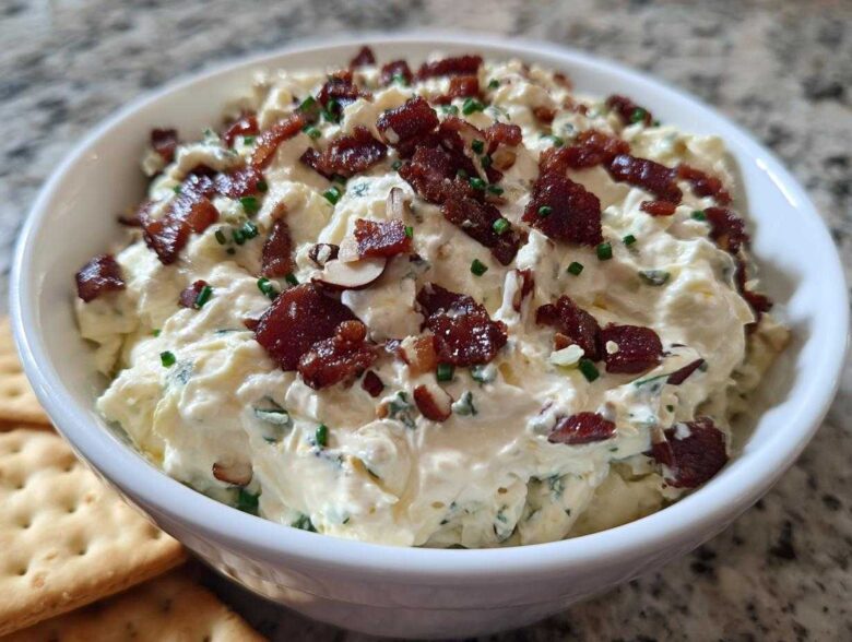 A close-up of a bowl filled with creamy Neiman Marcus dip, topped generously with crispy bacon pieces and chopped chives.