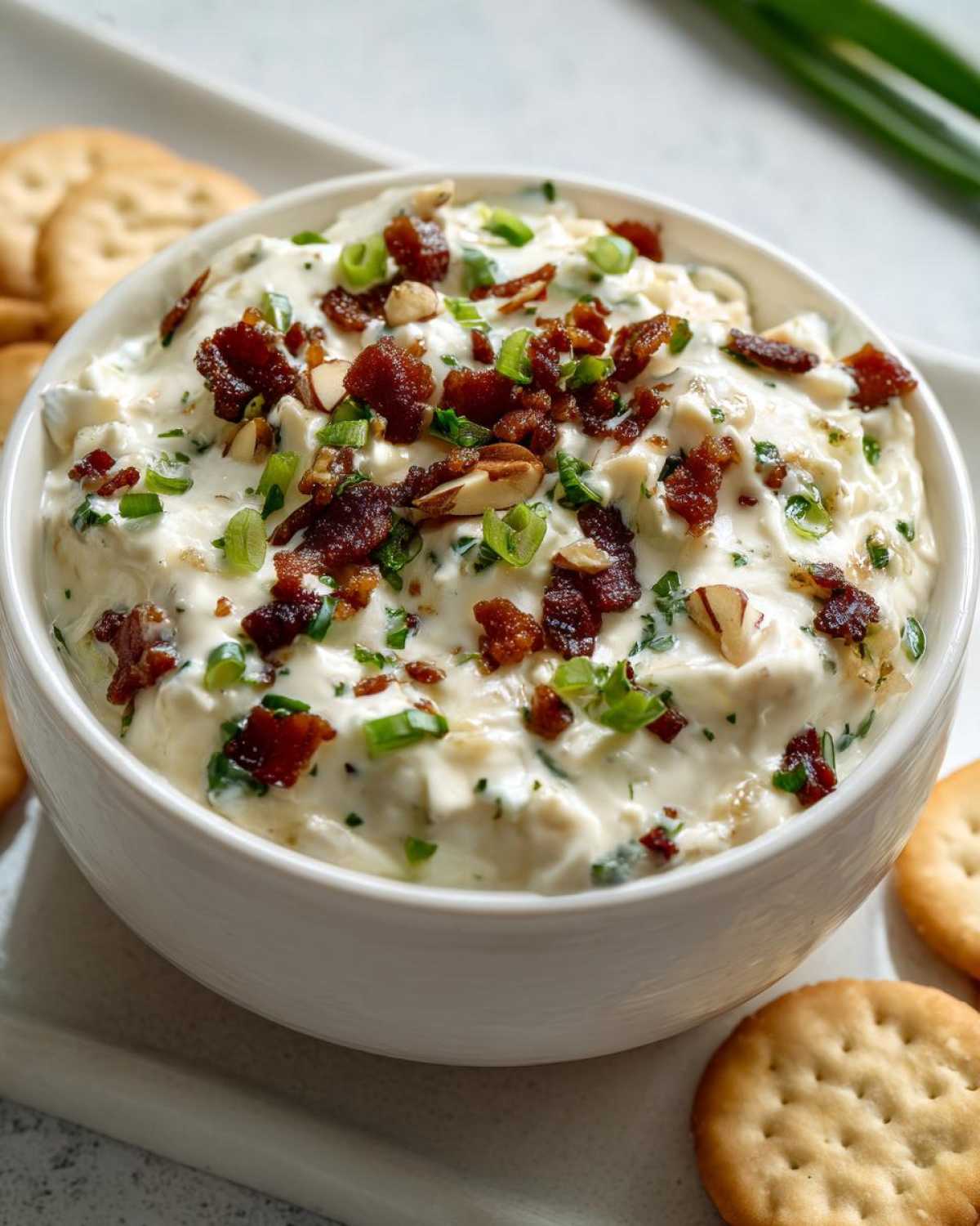 Close-up of creamy Neiman Marcus dip in a white bowl, topped with bacon bits, sliced almonds, and green onions.