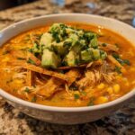 A close-up of a bowl of creamy tortilla soup garnished with shredded chicken, corn, avocado chunks, and crispy tortilla strips.