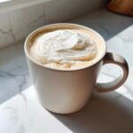 A creamy white hot chocolate topped with whipped cream in a light speckled mug.