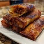A stack of golden brown, crispy egg roll appetizers, with one cut open to show the savory meat filling.