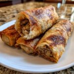 Close-up of three golden brown, crispy fried egg roll appetizers stacked on a white plate, showing savory filling.