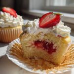 A cross-section view of a strawberry shortcake cupcake showing the filling, topped with whipped cream and a fresh strawberry slice.